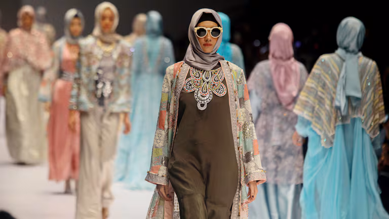 Model wearing a hijab, sunglasses, and an embellished coat walking a fashion show runway
