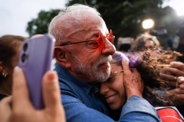 Luiz Inácio Lula da Silva wearing red star-shaped glasses while hugging a smiling supporter