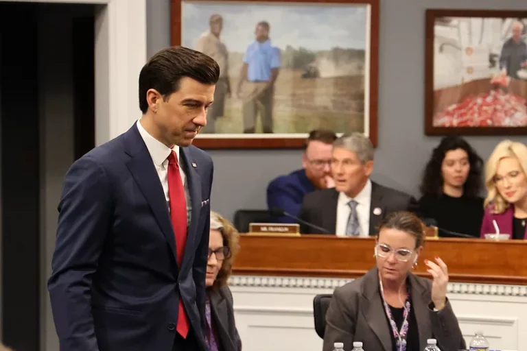 A man in a navy suit and red tie walking through a hearing room past seated committee members