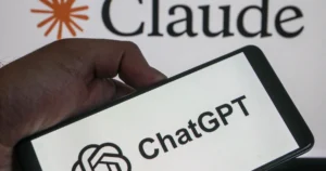 A hand holding a smartphone displaying the ChatGPT logo in front of a screen showing the Claude logo