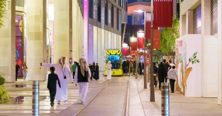 Pedestrians walk along a modern city street lined with Qatari flags as a bright yellow tram approaches