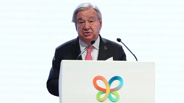 António Guterres speaking into microphones at a white podium with a colorful butterfly logo