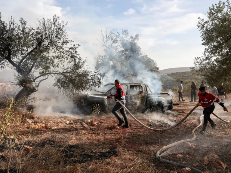 Emergency workers spray water onto a burning pickup truck emitting thick smoke in an olive grove