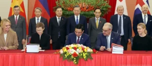 Officials sign documents during an official ceremony while standing dignitaries look on in the background
