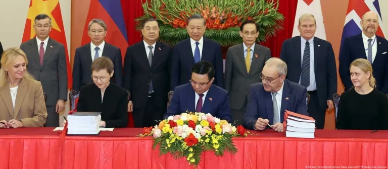 Officials sign documents during an official ceremony while standing dignitaries look on in the background