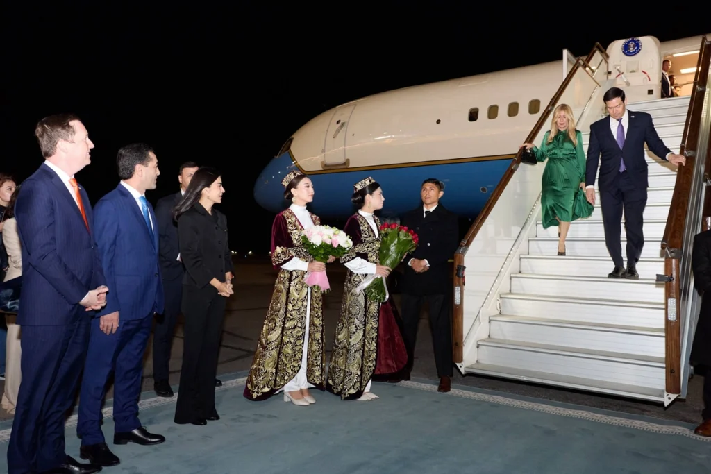 A diplomat and companion descend airplane stairs at night, greeted by women in ornate traditional attire holding flowers