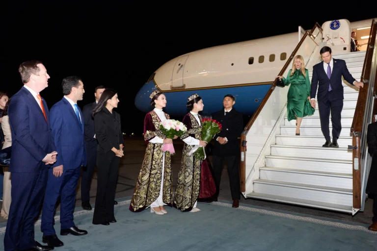 A diplomat and companion descend airplane stairs at night, greeted by women in ornate traditional attire holding flowers
