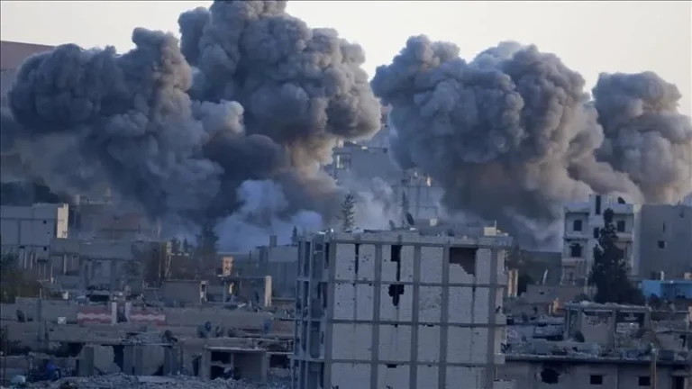 Massive plumes of thick grey smoke rise above damaged buildings in an urban area following an explosion