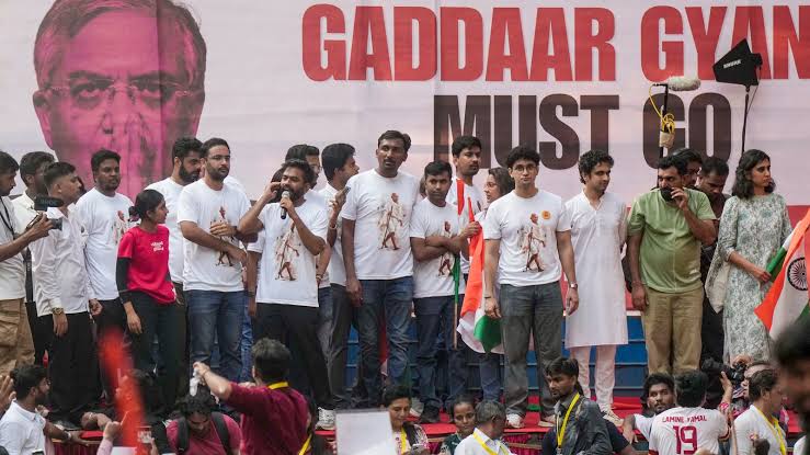 Protesters gathered on stage with a speaker in front of a banner reading GADDAAR GYAN MUST GO