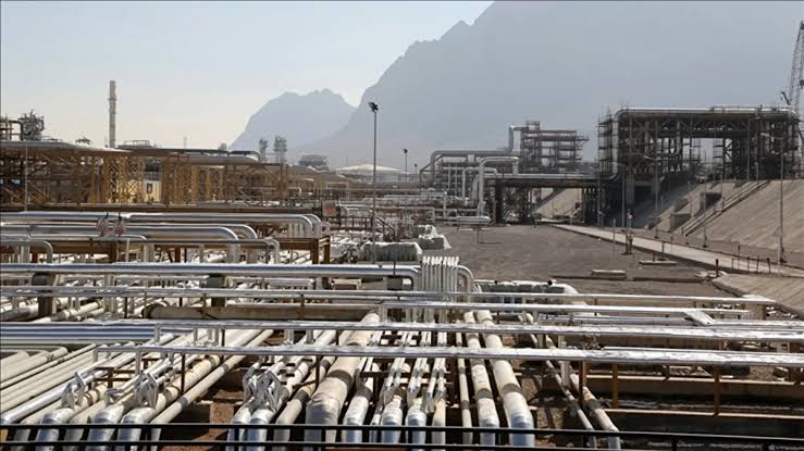 Industrial plant with complex networks of silver pipelines and steel structures set against mountains in the background