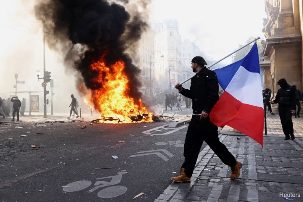A protester carrying a French flag walks past a large fire burning in a city street during a demonstration