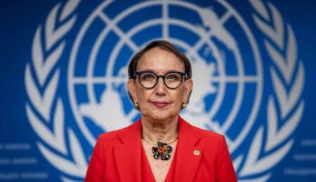 Rebeca Grynspan wearing a red blazer and glasses in front of the United Nations emblem