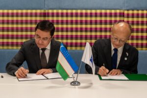 Two men in suits sign documents at a table with small desk flags, including the flag of Uzbekistan