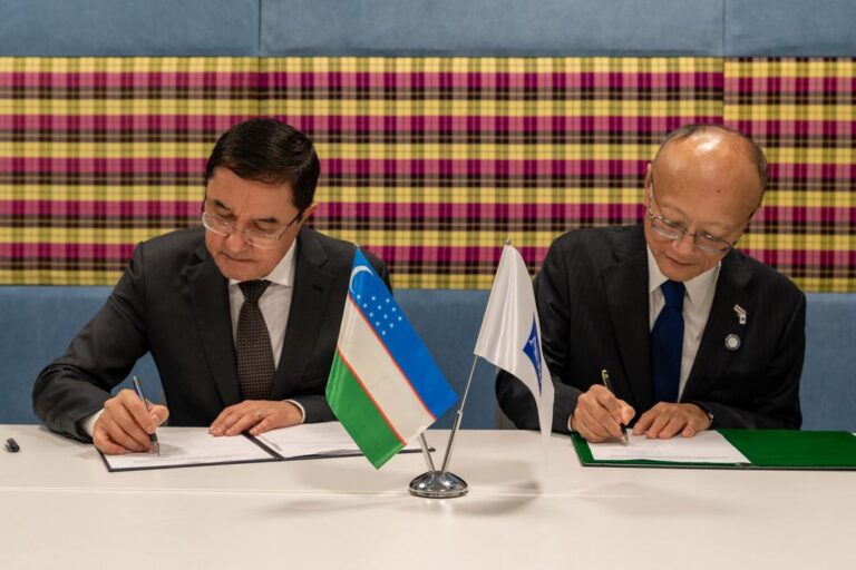 Two men in suits sign documents at a table with small desk flags, including the flag of Uzbekistan