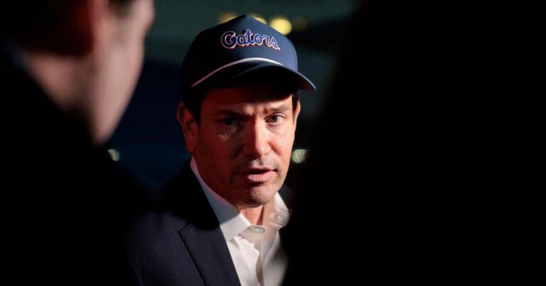 Ron DeSantis wearing a navy blue Gators baseball cap and suit jacket while speaking