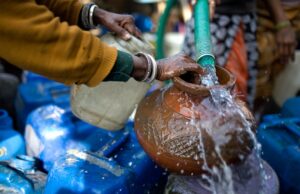 Hands guiding water from a hose into a clay pot surrounded by blue plastic containers