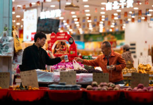 A shopper in an orange jacket buys fruit from a vendor at a brightly lit Chinese market