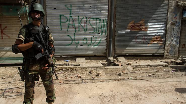 An armed soldier stands guard on a street in front of closed shop shutters with green graffiti reading Pakistan Zindabad