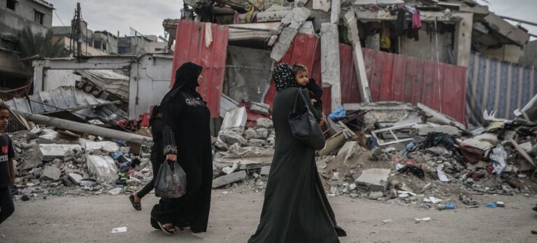 Two women wearing black abayas, one holding a young child, walk past destroyed buildings surrounded by rubble