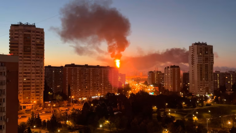 A large fiery explosion with a thick plume of dark smoke rises above apartment buildings in a city at dusk