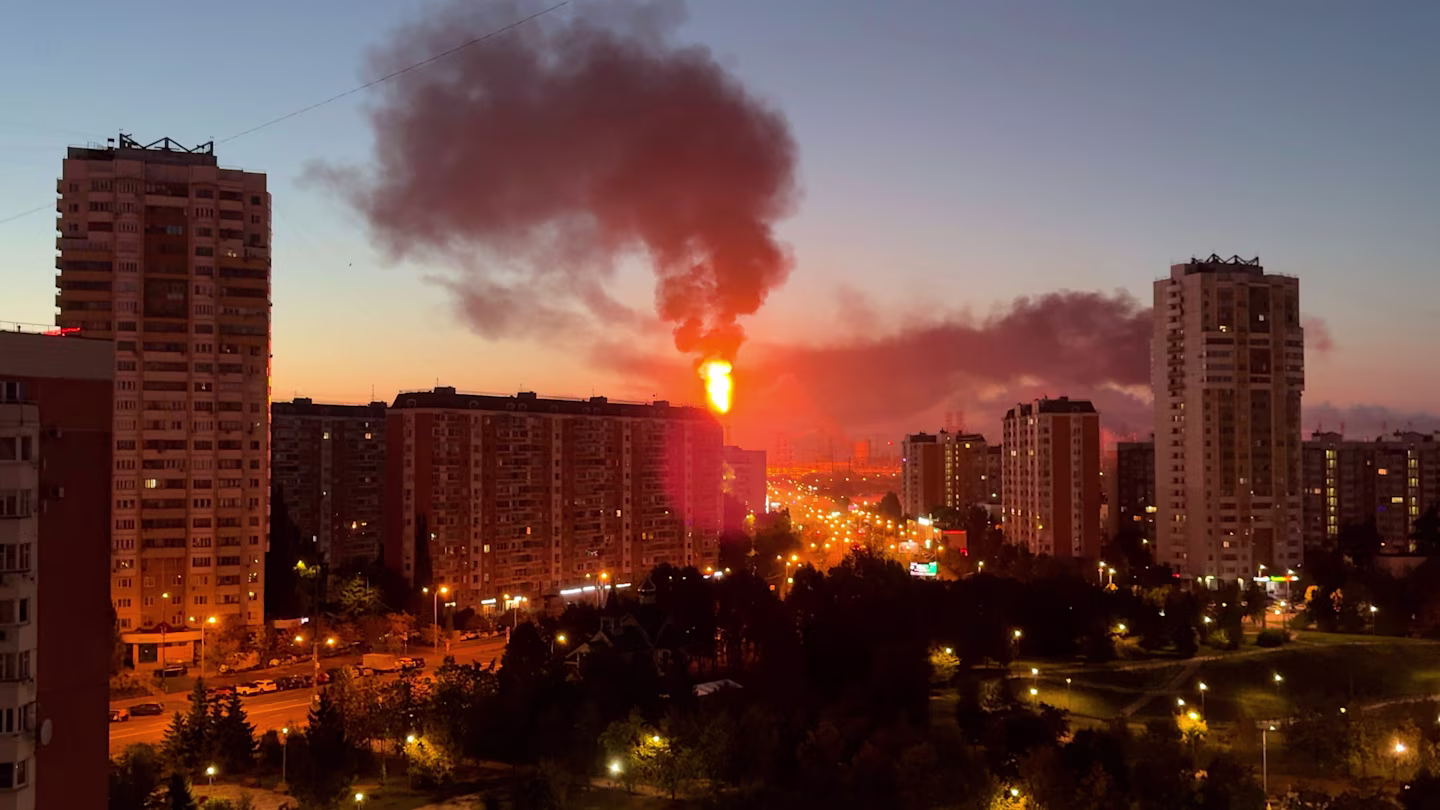 A large fiery explosion with a thick plume of dark smoke rises above apartment buildings in a city at dusk