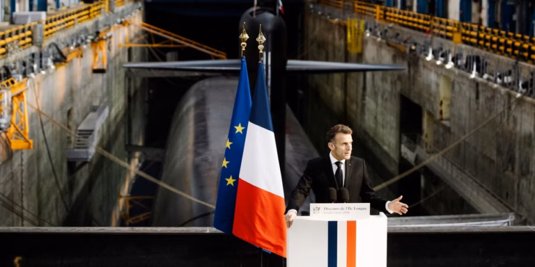 Emmanuel Macron speaking at a podium with French and EU flags, in front of a large submarine in a dry dock