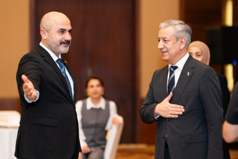 Two men in dark suits greet each other, one gesturing welcomingly while the other places his hand over his heart