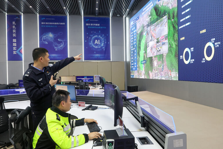 An officer points to a large screen displaying maps and camera feeds while another sits at a command center console
