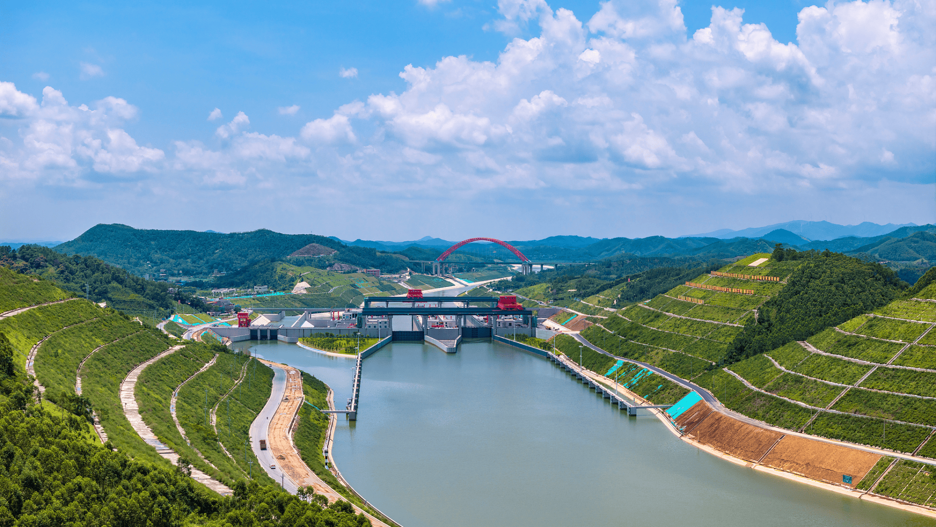 Aerial view of a canal lock system nestled between terraced green hills, with a red arch bridge in the distance