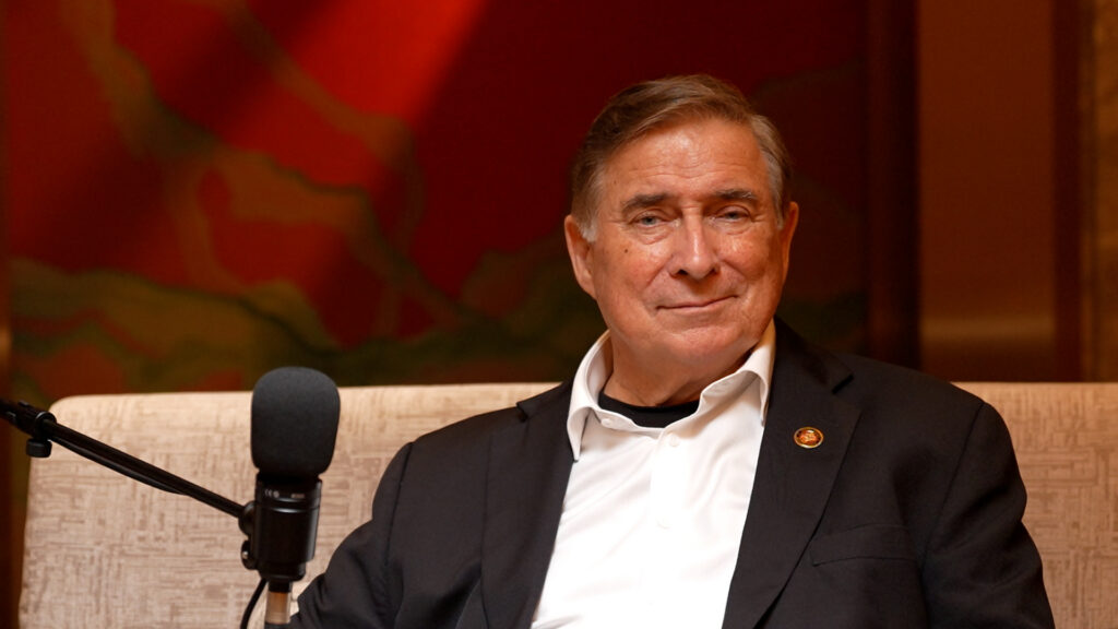 An older man wearing a dark suit jacket and white shirt smiling near a microphone