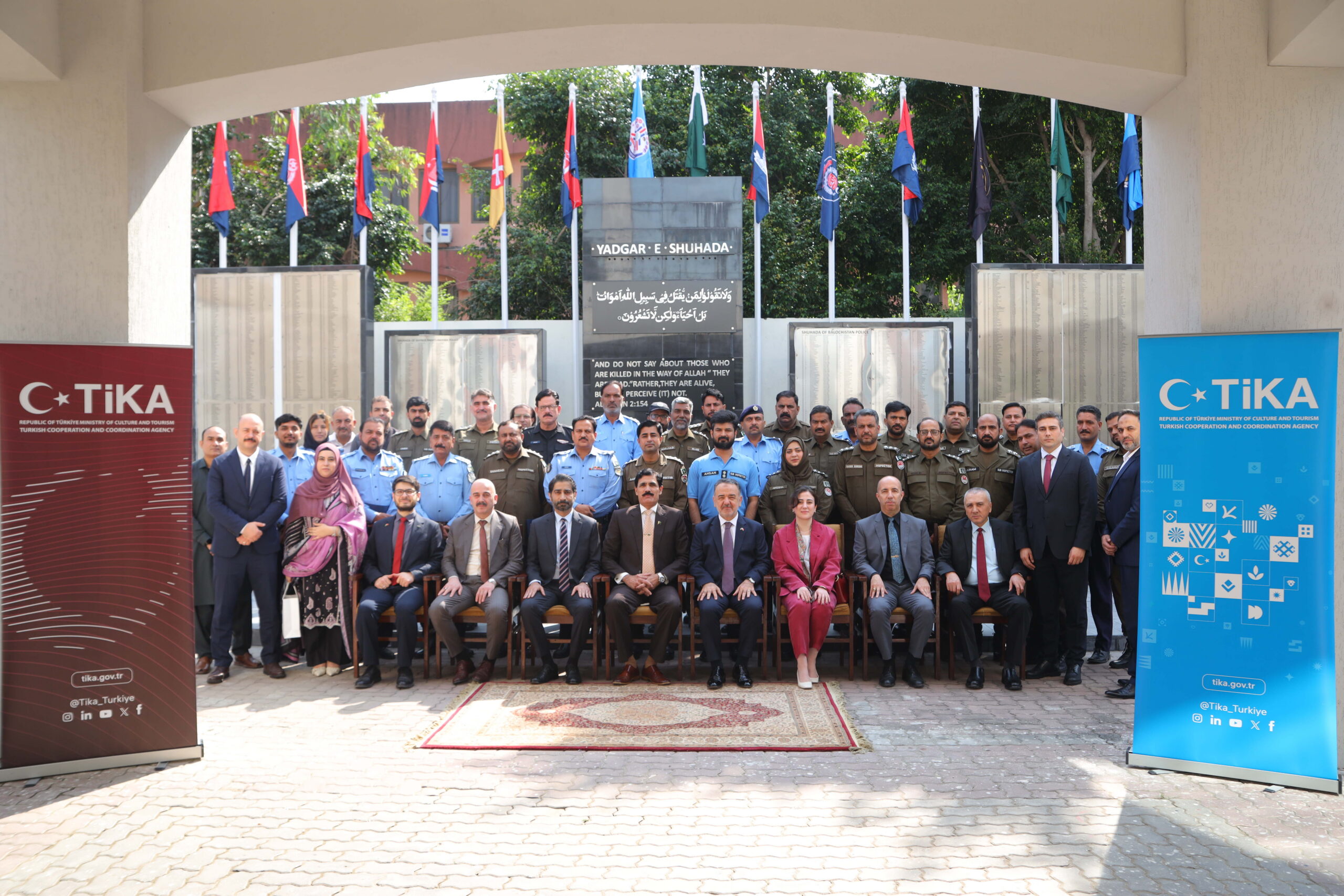 Officials and police officers posing in front of the Yadgar-e-Shuhada memorial, flanked by TIKA banners
