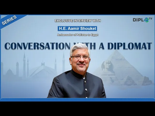 Promotional graphic for a series titled Conversation with a Diplomat featuring H. E. Aamir Shouket Ambassador of Pakistan to Egypt. A middle aged man with gray hair and glasses wearing a dark formal suit smiles in front of a blue background with faint images of pyramids and Faisal mosque silhouettes. Text on the image reads Exclusive Interview with H. E. Aamir Shouket Ambassador of Pakistan to Egypt Series and DIPLOTV.