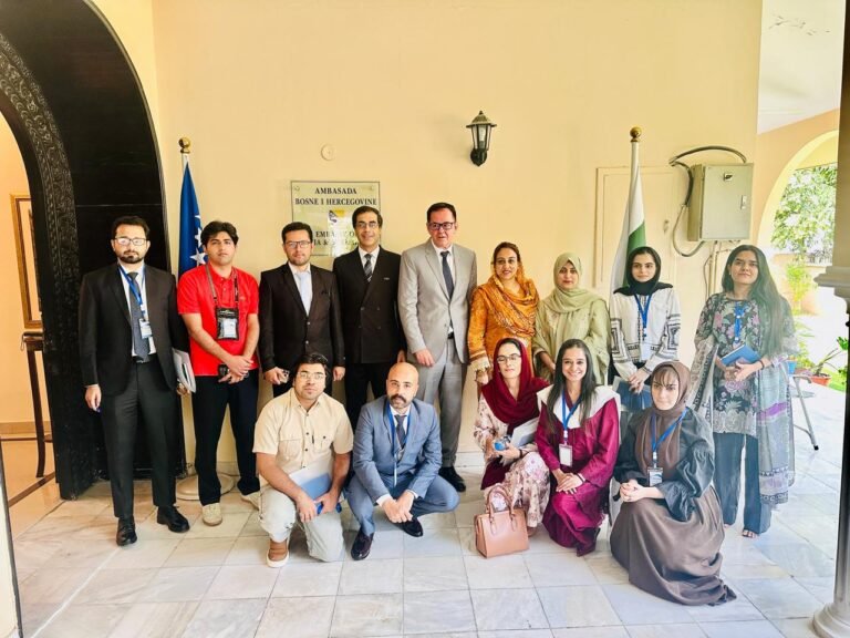 A group of fifteen people posing for a formal photo at the entrance of the Embassy of Bosnia and Herzegovina. The group consists of men and women in professional attire, including suits and traditional Pakistani wear; several members are wearing blue lanyards. In the background, a plaque identifies the embassy, and the national flags of Bosnia and Herzegovina and Pakistan are displayed on either side of the group. The setting features a light-colored arched wall and a tiled floor.