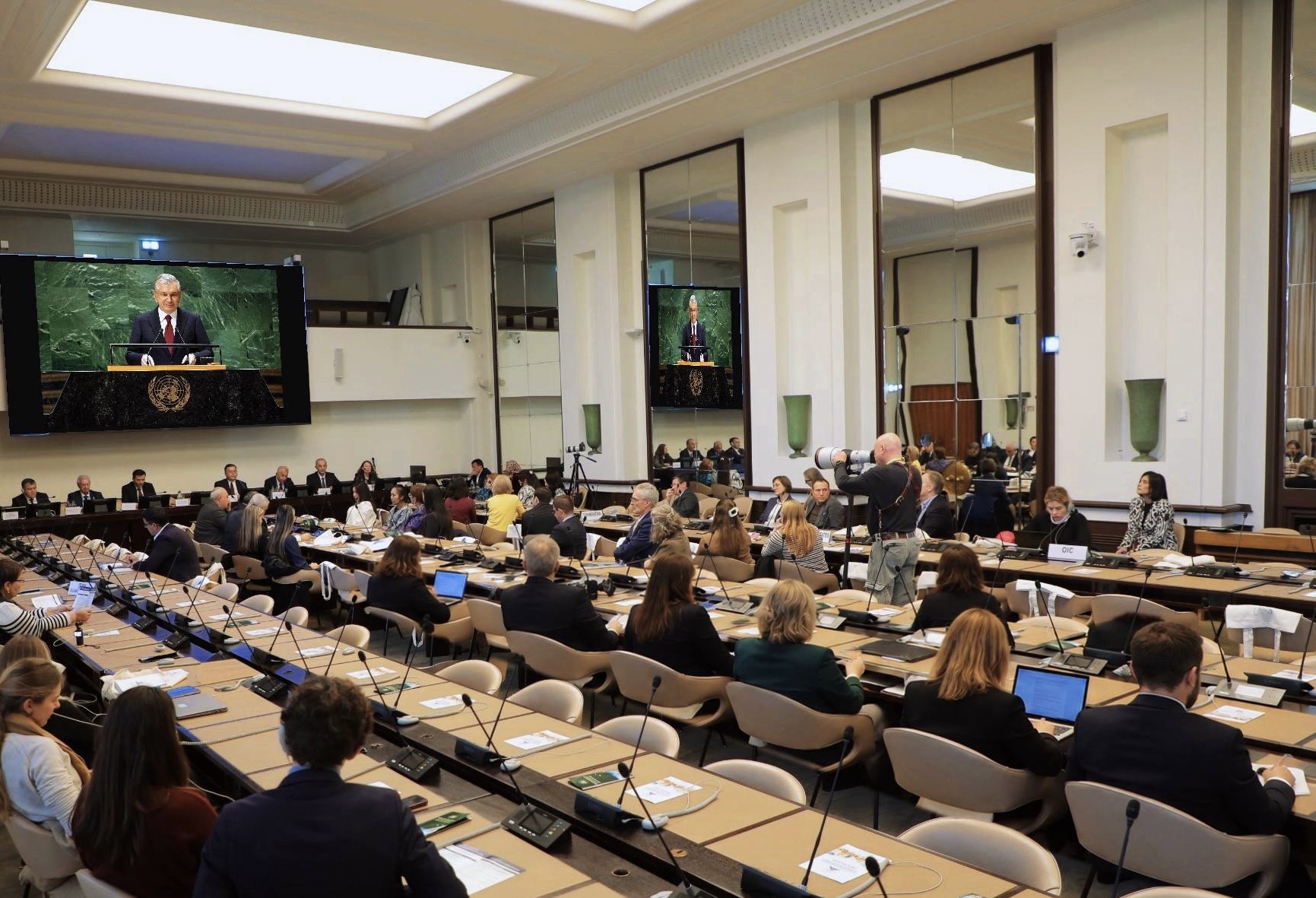 Delegates seated in a large United Nations conference room watching a speaker broadcast on a screen