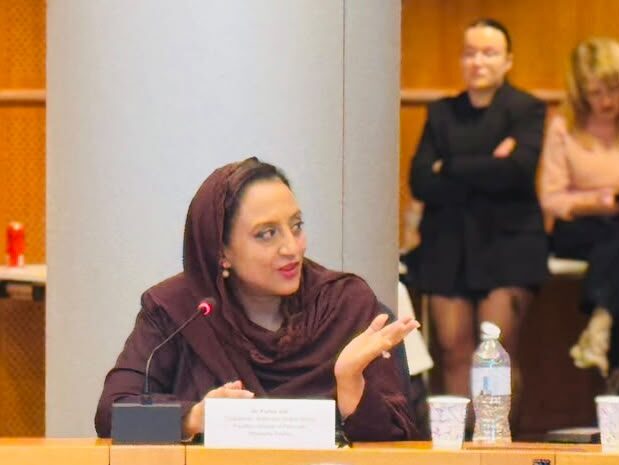 A woman wearing a brown headscarf gestures while speaking at a microphone during a conference panel